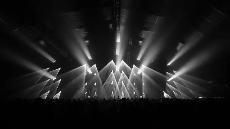 High-contrast view of a concert stage with massive light beams projecting from a central LED setup into the audience.